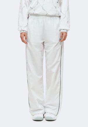 Personne portant un pantalon de survêtement blanc ample avec des bandes noires sur les côtés et des baskets blanches, debout devant un fond uni.