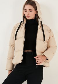 Beige puffer jacket with a hood, black drawstrings, and a full zipper. Features a quilted design and is paired with a black ribbed turtleneck.