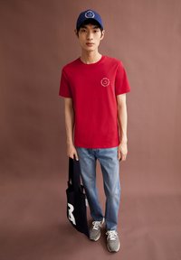 T-shirt en coton rouge avec un petit graphique blanc, casquette bleue avec logo, jean bleu, sac fourre-tout noir avec lettrage blanc, baskets grises.