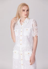 Blouse en dentelle blanche à manches courtes, dotée de deux poches avant et de boutons dorés. La dentelle présente un motif floral et une texture légère.
