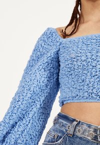Modrá, texturovaná blůza s nabranou látkou, off-shoulder design a vypufkovanými rukávy. Sestaveno s denimovými šortkami s vysokým pasem a průhledným páskem.