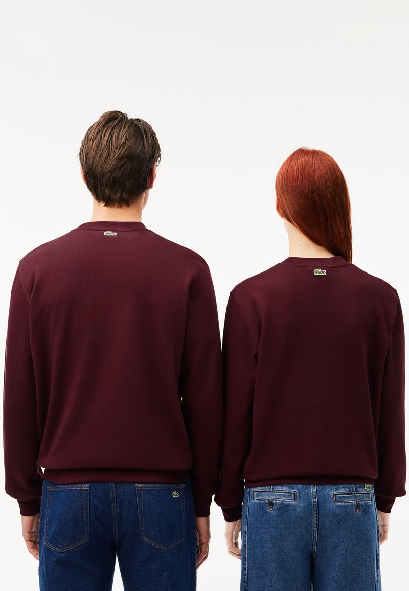 Sweatshirts bordeaux à manches longues et col rond, avec un petit logo Lacoste vert à l'arrière. Jeans bleus visibles sous la taille.