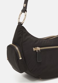 Černá nylonová taška přes rameno se zlatými kovovými doplňky, s několika kapsami na zip a zaobleným tvarem. Měkká textura a nastavitelný popruh.