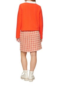 Pull en maille matelassée orange avec col blanc, associé à une jupe vichy rouge et blanche. Le mannequin porte des bottes blanches à talons chunky.