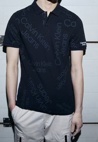 Man som bär en svart polo skjorta från Calvin Klein Jeans med upprepat varumärke, i kombination med ljusa cargobyxor, med händerna avslappnat vid sidorna.