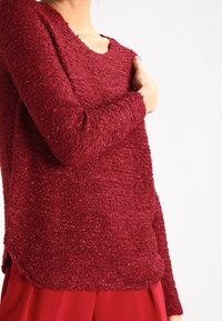 Maglione rosso a trama con maniche lunghe, caratterizzato da una vestibilità leggermente ampia e un ampio scollo. Il tessuto appare morbido e peloso.