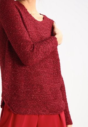 Maglione rosso a trama con maniche lunghe, caratterizzato da una vestibilità leggermente ampia e un ampio scollo. Il tessuto appare morbido e peloso.