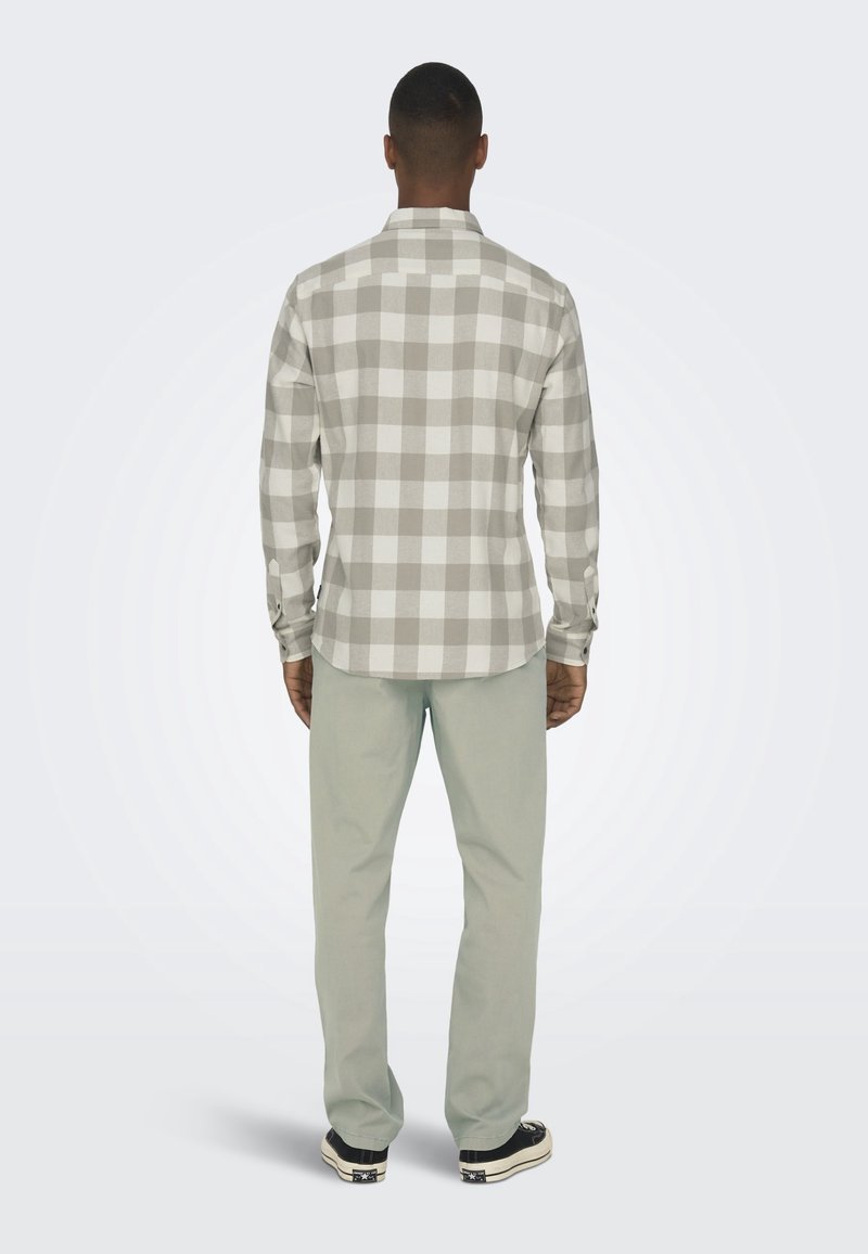 Homme vu de dos portant une chemise à manches longues beige et blanche à carreaux, un pantalon gris clair et des baskets noires et blanches.