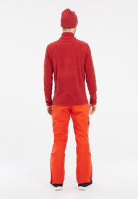 Rode fleece longsleeve top met hoge kraag gecombineerd met feloranje sneeuwbroek met zijritsen. De outfit wordt compleet gemaakt met een rode beanie.