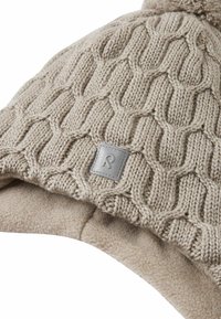 Gebreide beige beanie met een gestructureerd patroon, een grijze leren logo patch en een zachte fleece voering voor extra warmte. Afgeronde bovenkant met pompon.
