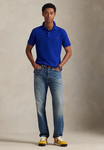 Fiatal férfi áll zsebre dugott kézzel, kék pólót, kifakult farmert, sárga sportcipőt és karórát visel semleges háttér előtt.