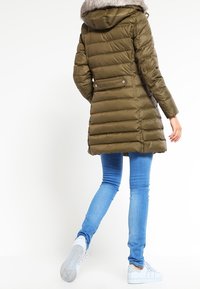 Kvinna klädd i en olivgrön vadderad jacka med huva i päls, ljusblå jeans och vita sneakers som går bort från en vit bakgrund.