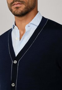 Uomo che indossa una camicia a righe blu chiaro sotto un cardigan scuro blu navy con rifiniture e bottoni bianchi, tagliato all'altezza del mento e delle spalle.