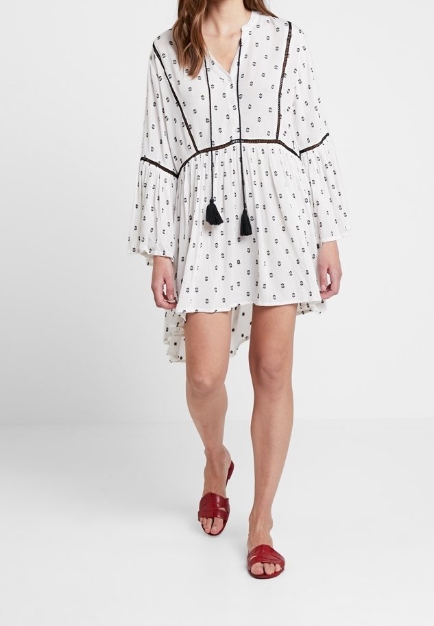 Femme portant une robe blanche à manches longues avec des motifs noirs et des franges, associée à des sandales plates rouges à bout ouvert, marchant vers l'avant.