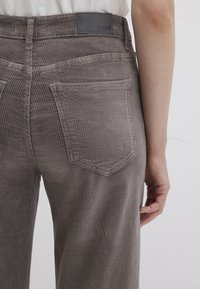 Gray corduroy pants with a textured surface, featuring two back pockets and a logo patch at the waistband. Visible vertical ribbing detail.