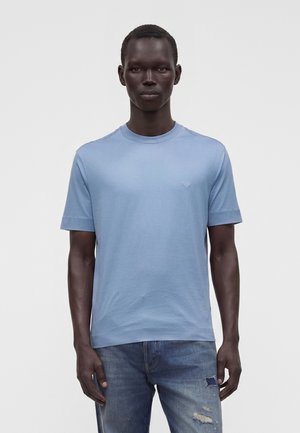 Bărbat îmbrăcat cu un tricou bleu cu mâneci scurte și blugi albaștri deteriorați, stând în fața unui fundal simplu și deschis la culoare.