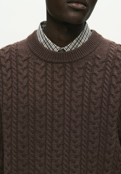 Rjav pulover z kabelskim pletenjem in okroglo ovratnikom, nosen čez belo in modro karirasto srajco. Tekstura se zdi mehka in prijetna.