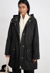 Zwarte waterdichte parka met gouden accenten, uitgerust met een verstelbare taille, grote voorzakken en een groene plaidvoering in de capuchon.