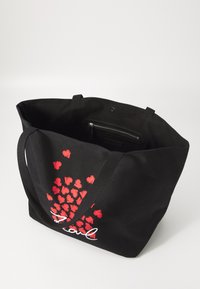 Sac en toile noir avec un motif de cœurs rouges et du texte blanc, doté de deux bretelles et d'une poche zippée à l'intérieur.