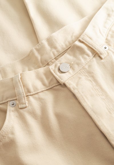 Zbliżenie na beżowe jeansy z denimu pokazujące pas, szlufki na pasek, metalowy guzik z grawerem "W.W." oraz nity przy kieszeni.