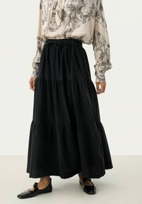 Gonna maxi nera a strati con vita elastica, caratterizzata da una texture liscia e una forma fluida, abbinata a una blusa a fantasia e scarpe basse.