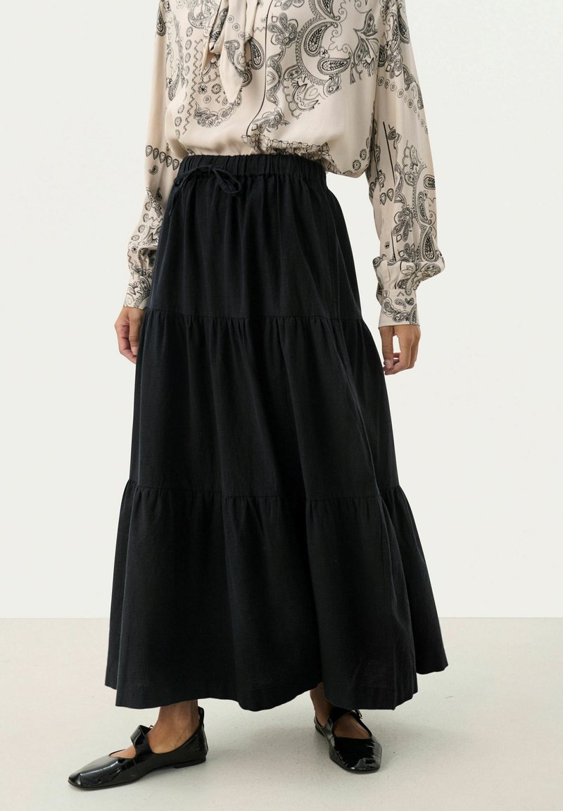 Gonna maxi nera a strati con vita elastica, caratterizzata da una texture liscia e una forma fluida, abbinata a una blusa a fantasia e scarpe basse.