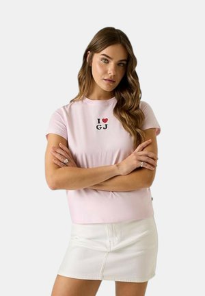 Donna con lunghi capelli castani che indossa una t-shirt rosa chiaro con il testo "I love GJ" e una minigonna bianca, in piedi con le braccia incrociate.