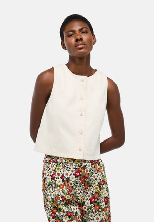 Femme portant un haut sans manches écru à boutons et un pantalon taille haute à motifs floraux orange, rouges, jaunes et blancs sur fond noir.