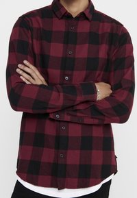 Chemise à carreaux rouge et noir à boutons avec des manches longues, en flanelle, présentant un ourlet arrondi et des boutons noirs.