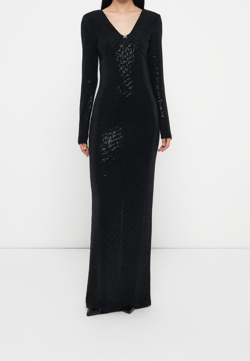 Femme portant une longue robe en maille noire à manches longues et col en V, debout contre un mur clair uni.