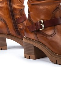 Botas de cuero marrón hasta el tobillo con un acabado texturizado, cremallera lateral y una correa marrón con una hebilla de plata. Tacón de goma grueso y con patrón.