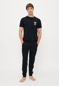 T-shirt preta de algodão com um gráfico de urso no peito, combinada com calças de moletom pretas com detalhes laterais. Modelo está descalço.
