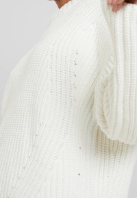 Maglione bianco chunky lavorato a maglia con una texture a coste, caratterizzato da un collo rotondo e dettagli di cucitura visibili sulle maniche.