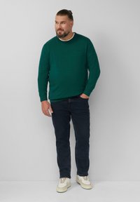 Groene gebreide trui met een ronde hals, gecombineerd met donkerblauwe jeans en witte sneakers. De stof lijkt glad en texturering te hebben.