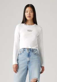 Valge pikka varrukat omaksvõtte särk, millel on hõbedane "Levi's" logo rinnal, koos kõrge vöökohtadega helesiniste kulunud teksadega.