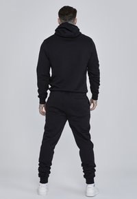 Black cotton hoodie with a large hood, paired with matching joggers. Joggers feature a pocket and ribbed cuffs; smooth texture throughout.