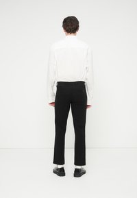 Pantalon noir à coupe ajustée, fabriqué dans un tissu lisse. Comprend des poches arrière et une ceinture standard, porté avec une chemise blanche.
