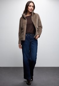 Brown faux fur jacket with a stand-up collar, worn over a dark brown top, paired with dark blue wide-leg jeans and brown footwear.