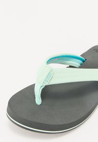 Mint green flip-flop with a textured strap and a black rubber sole, featuring a white outline. Brand logo visible on strap.
