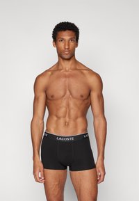 Sorte boxer-briefer med en hvid Lacoste-logo på taljen, lavet af et glat og strækbart stof. Tætsiddende design, midt på låret.