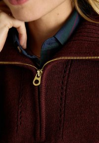 Burgunderfarbener Strickpullover mit einem goldenen Reißverschluss, der über feine Stickmuster verfügt. Darunter trägt er ein dunkles karierte Hemd mit Kragen.