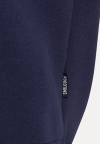 Fondo blu navy di un maglione con una texture liscia. Presenta un'etichetta con il logo "SMILODON" vicino al polsino. Design semplice, senza motivi o dettagli.
