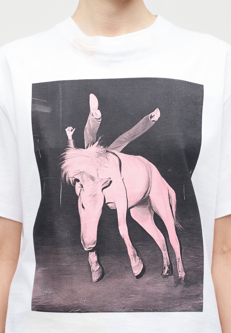 Persona che indossa una maglietta bianca con una grafica nero-rosa di un acrobata capovolto in equilibrio sulla schiena di un cavallo.