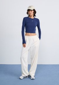 Kék hosszú ujjú crop top kis logóval, fehér, laza nadrágok és világos színű sportcipők. Világos háttér és kék padló.