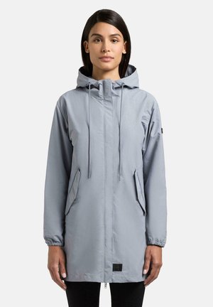 Frau trägt eine lange hellgraue Kapuzenjacke mit Fronttaschen und elastischen Bündchen, steht vor einem schlichten weißen Hintergrund.