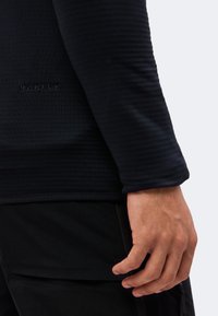 Gros plan d'une main reposant à côté d'un haut à manches longues noir texturé avec un logo Oakley subtil et un pantalon noir avec poche zippée.