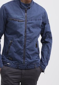 Marinblå jacka med hög krage, framsida med dragkedja och flera fickor med dragkedja. Har en slät textur och justerbar midja.