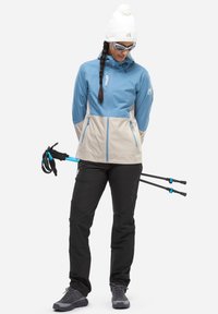 Helles blaues und beiges wasserdichtes Jackett mit Kapuze, schwarze Hose und graue Wanderschuhe. Inklusive Trekkingstöcke mit blauen Akzenten.