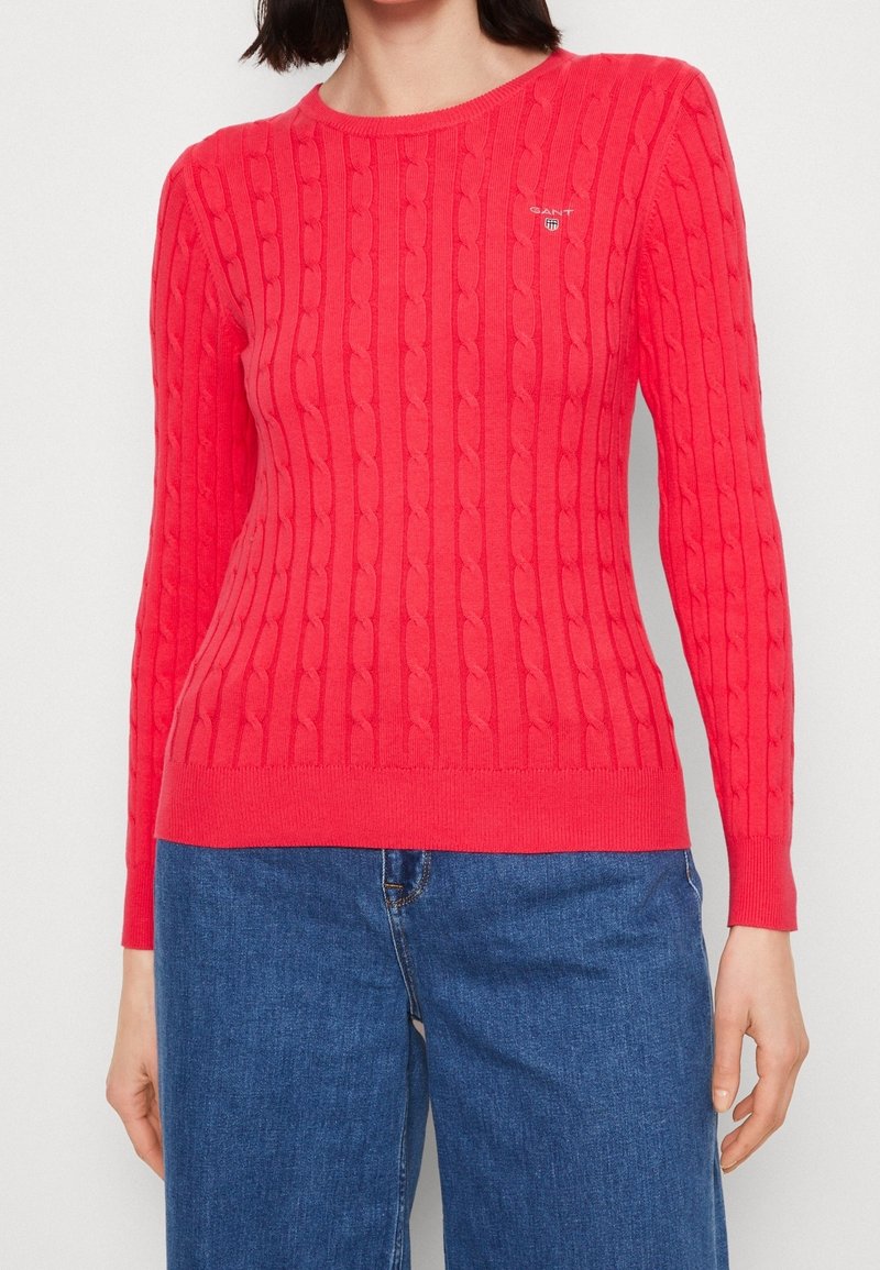 Kvinde iført en tætsiddende, knaldrød kabelstrikket sweater med lange ærmer og blå denimjeans, hænder afslappet langs siden.