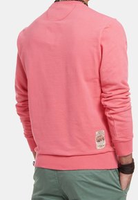 Růžová mikina s žebrovaným límcem a manžetami. Na spodním lemu má malý štítek. Materiál vypadá měkký a texturovaný.